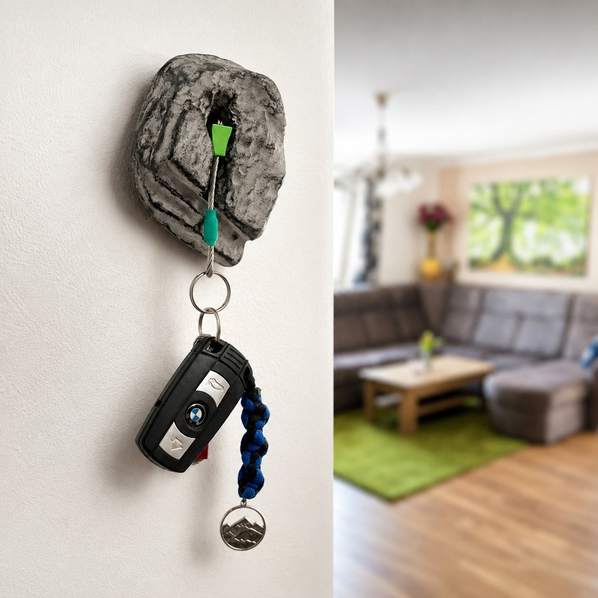 A novelty keychain holder shaped like a gray rock mount on a white wall, holding a black car key fob and blue beaded keychain. The right side shows a blurred living room interior.