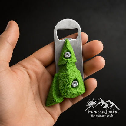 A hand holding a green climbing hold bottle opener with a textured surface and brushed metal blade against a dark background.