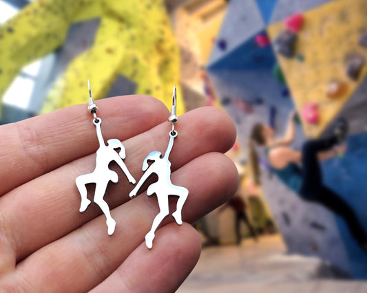 Silver climbing earrings held in a hand with a climbing wall in the background