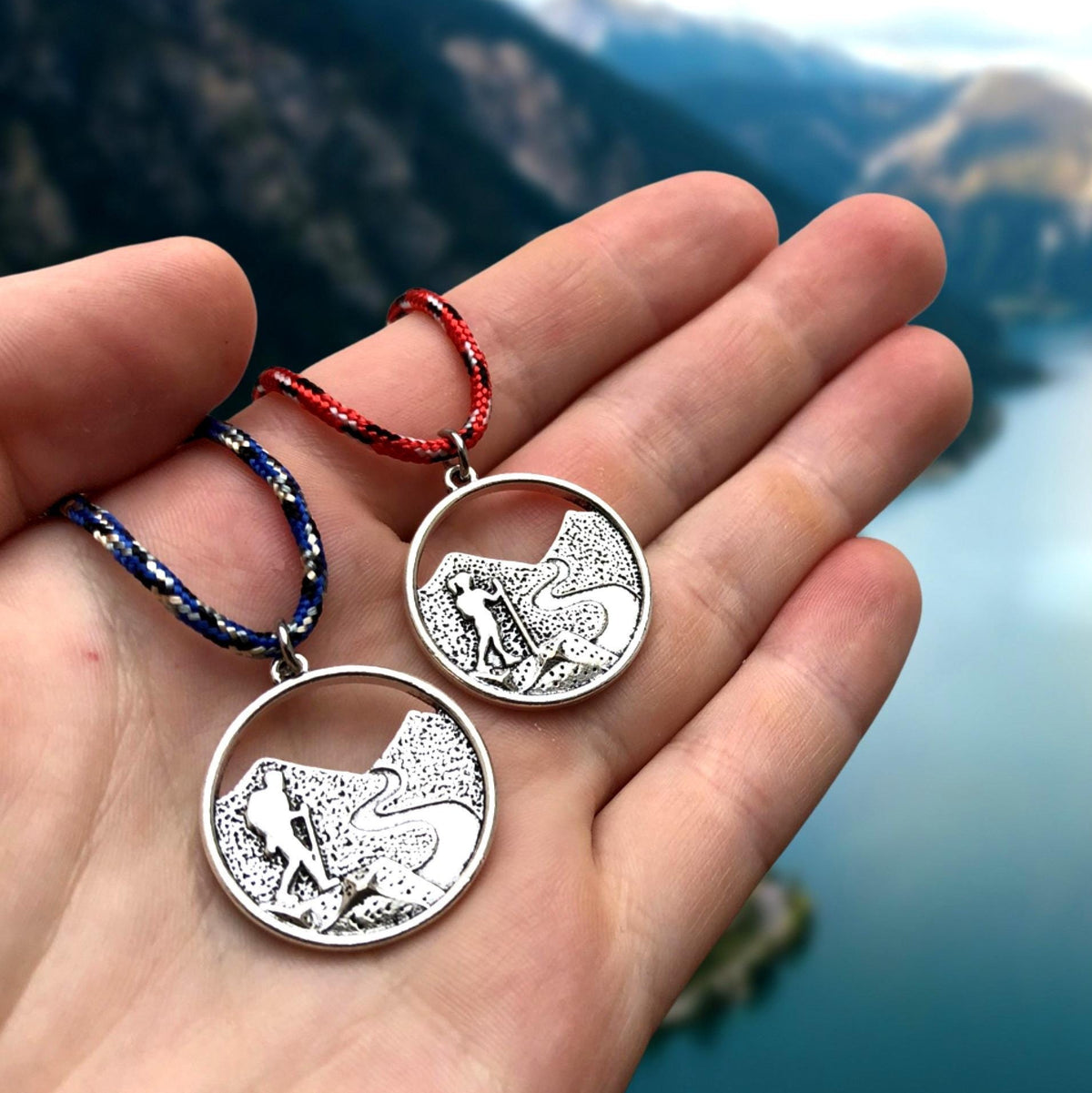 Two silver pendants with mountain designs held in a hand against a mountainous landscape.