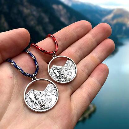 Two silver pendants with mountain designs held in a hand against a mountainous landscape.