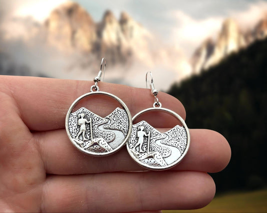 Silver earrings with Hiking designs held against a blurred natural background