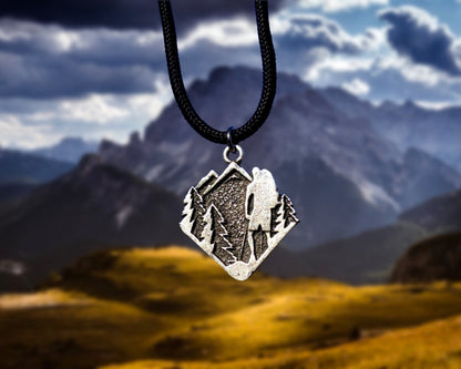 Silver pendant with mountain design on a black cord against a mountainous landscape