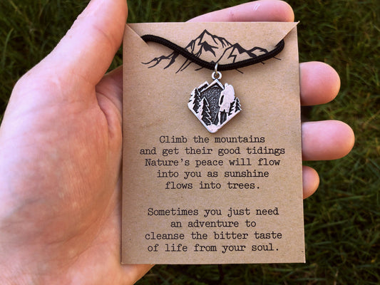Hand holding a necklace with a mountain pendant on a brown card with text against a grassy background