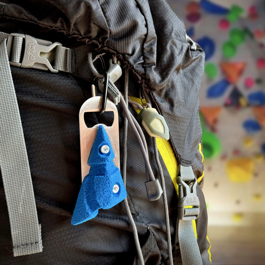 A blue climbing hold bottle opener attached via carabiner to a climbing harness for easy outdoor carrying.