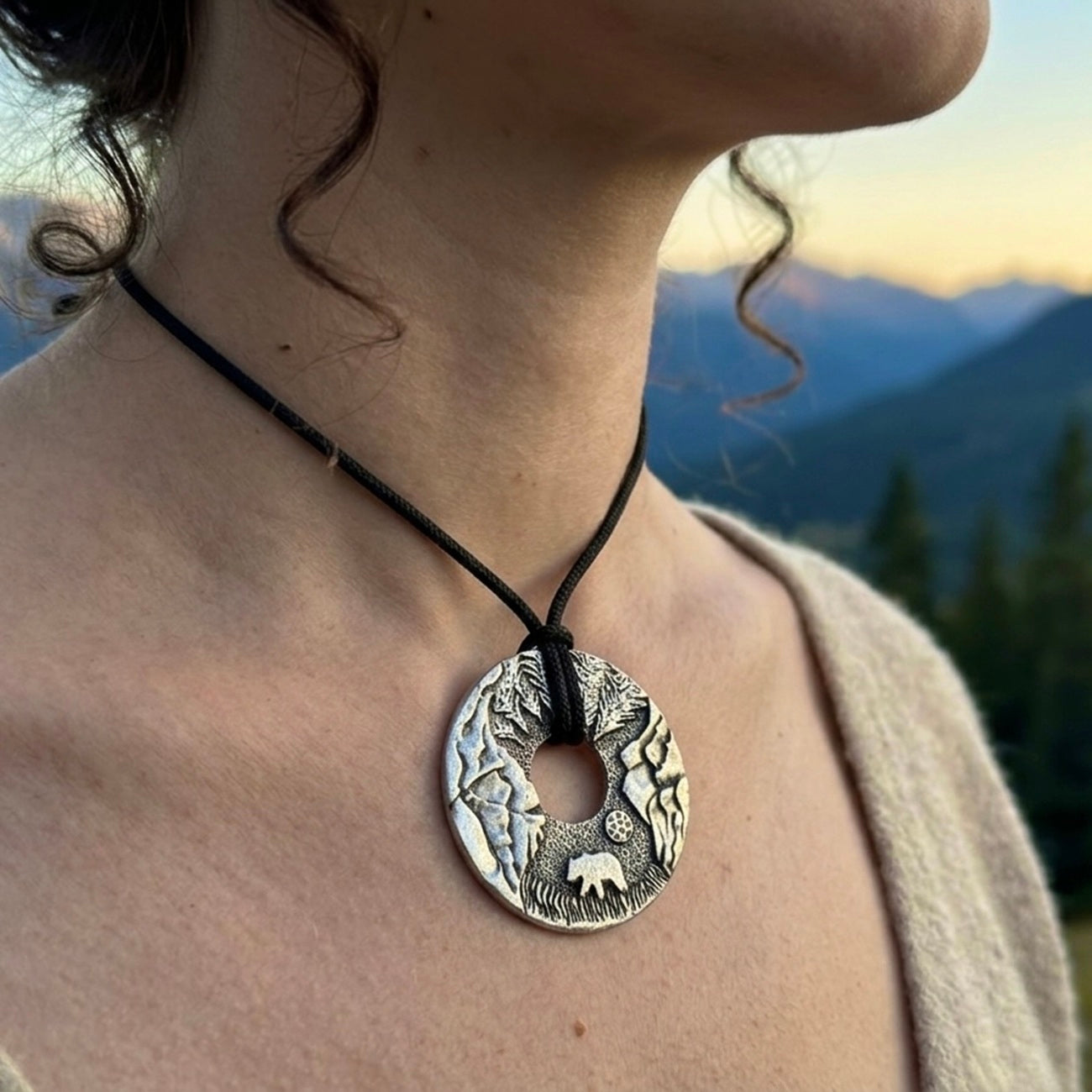 Person wearing a silver necklace with a nature-themed pendant against a mountainous background