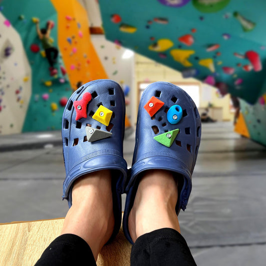 Blue clogs with colorful buttons worn by a person in front of a climbing wall.