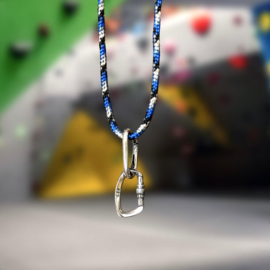 A silver openable carabiner necklace with blue and black accents, hanging against a climbing wall with various climbing hold accessories.