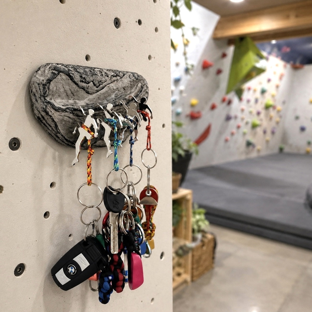 A wall-mounted climbing-peg key holder shaped like a rock, shown attached to a beige climbing wall in an indoor climbing gym. Several keys and keychains are hanging from the pegs, with a blurred climbing wall and padded floor visible in the background.