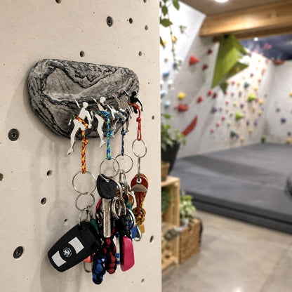 A wall-mounted climbing-peg key holder shaped like a rock, shown attached to a beige climbing wall in an indoor climbing gym. Several keys and keychains are hanging from the pegs, with a blurred climbing wall and padded floor visible in the background.