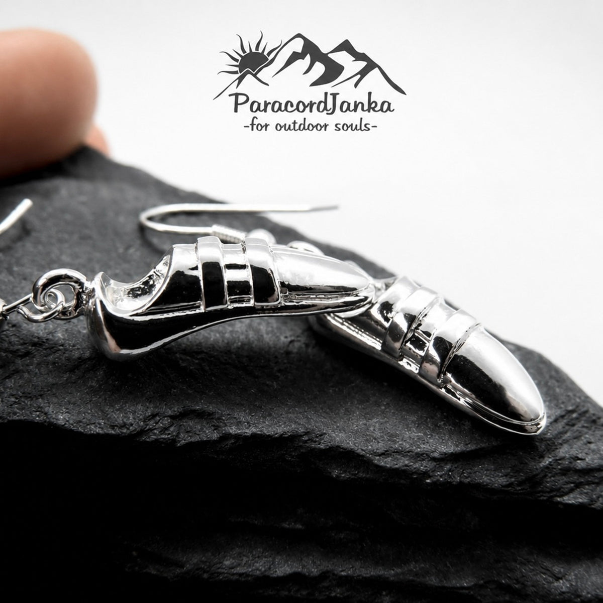 Silver-tone shoe-shaped dangle earrings displayed on a dark slate surface, with a finger partially visible at the top left and the ParacordJanka logo at the top center.