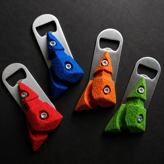 Four climbing hold bottle opener keychains in red, blue, orange, and green, arranged on a dark textured background. Each opener features a textured faux-leather Christmas tree design attached to a brushed metal keychain blade with a silver screw accent.