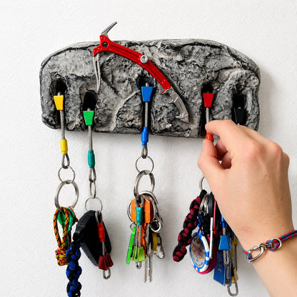 A textured gray rock-shaped wall key holder with multiple colored hooks, shown mounted on a white wall. A hand is reaching in to insert a key, and climbing-related keys and cords are hanging from the hooks.