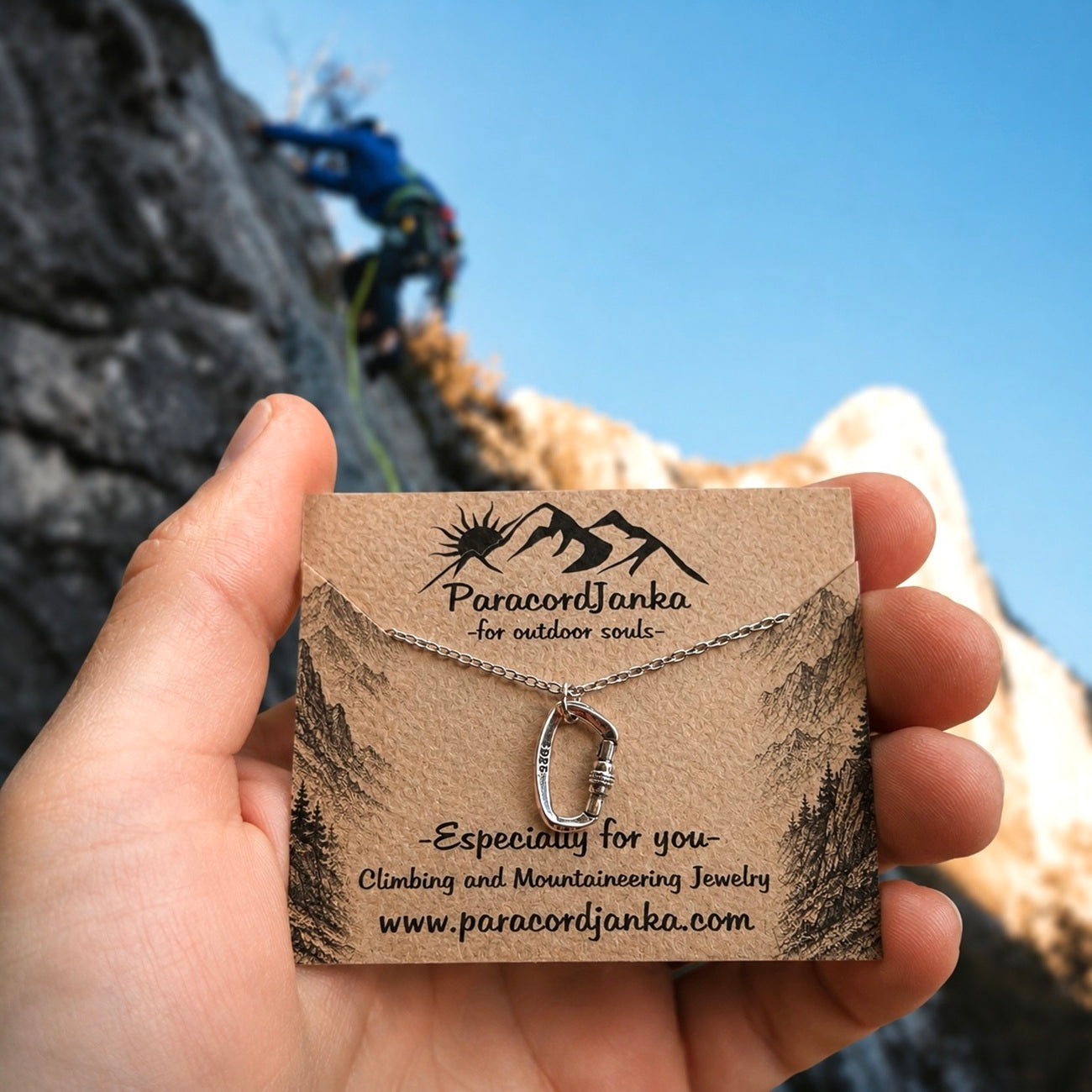 A hand holds a silver climbing loop necklace on a brown kraft display card. The background shows a blurred outdoor mountain climbing scene with a climber and blue sky.