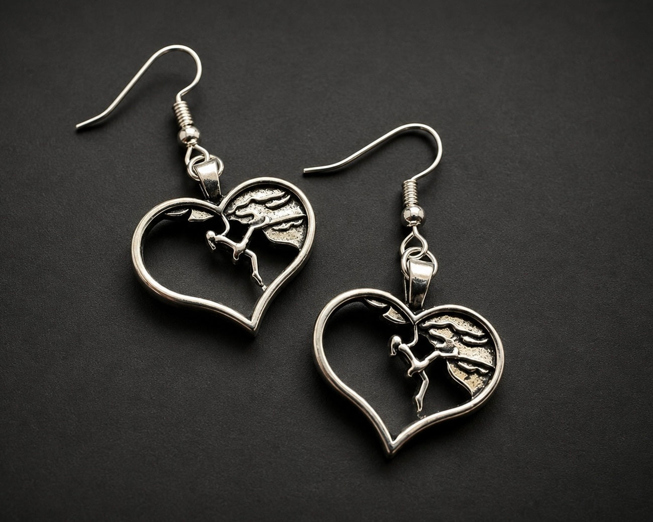 A pair of silver-toned drop earrings featuring heart-shaped cutouts with a detailed horse and rider motif in the center, displayed on a dark textured background.