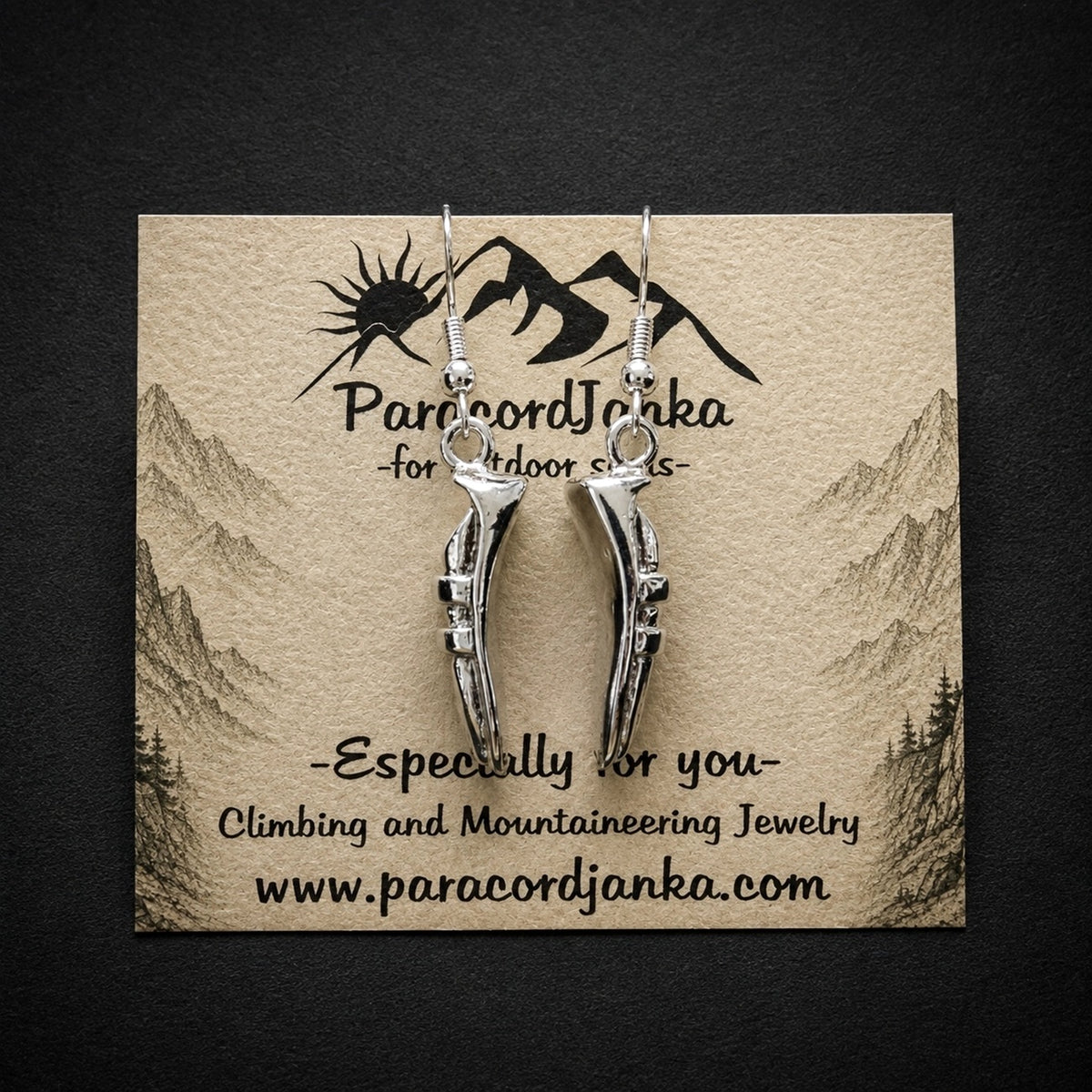Silver mountain-shaped dangle earrings displayed on a kraft paper card with the brand name 'ParacordJanka' and the website printed on it, set against a dark textured background.