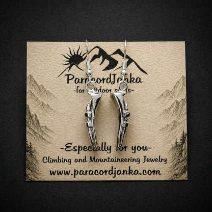 Silver mountain-shaped dangle earrings displayed on a kraft paper card with the brand name 'ParacordJanka' and the website printed on it, set against a dark textured background.