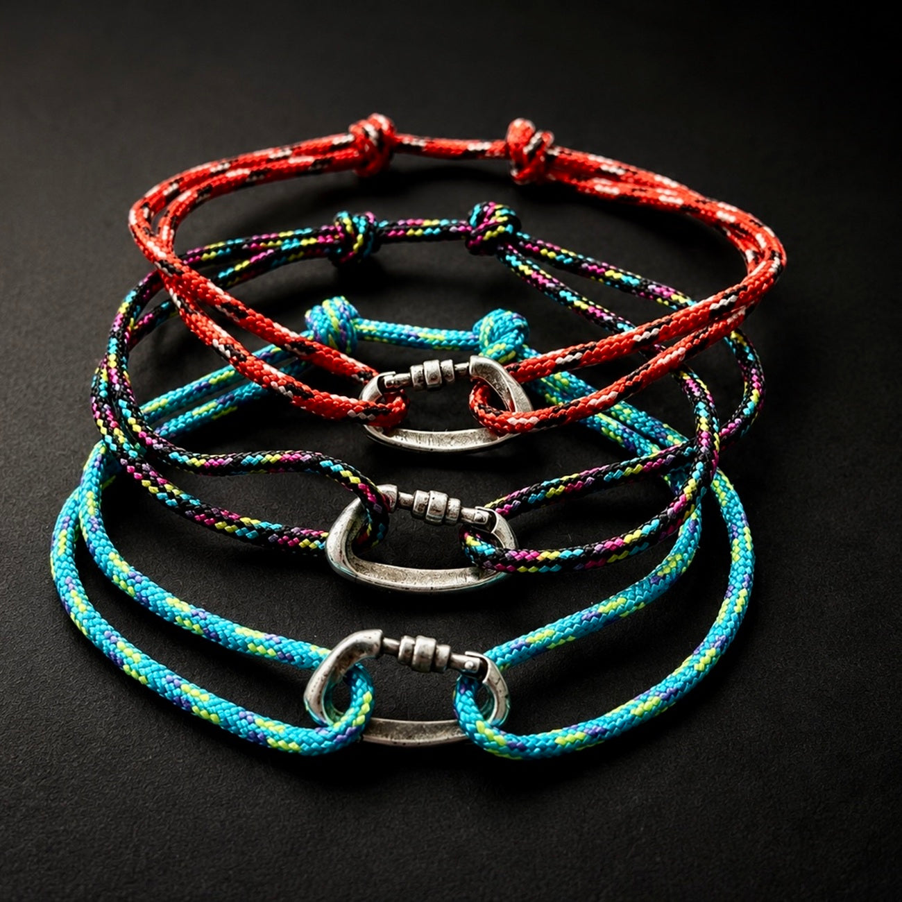 Four braided cord bracelets with silver-toned carabiner-style clasps, arranged in a stack on a dark background. The cords are multicolored, featuring red, blue, green, and purple threads.