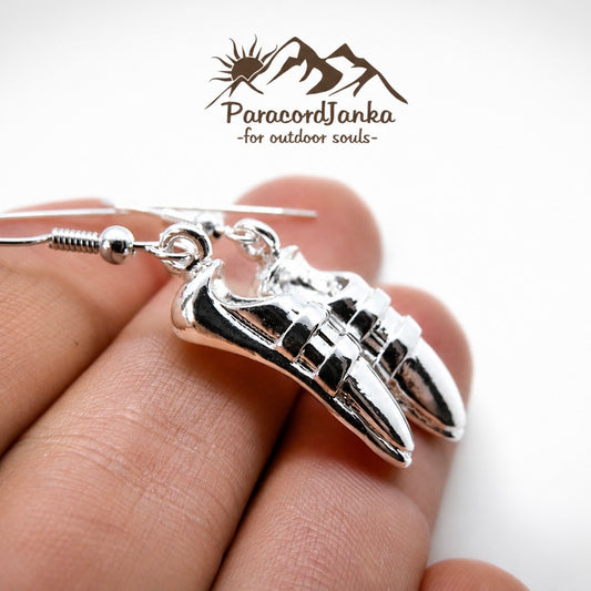 A pair of silver dangling earrings shaped like athletic sneakers, shown being held on a person's fingers against a white background. The brand logo 'ParacordJanka -for outdoor souls-' is visible at the top.