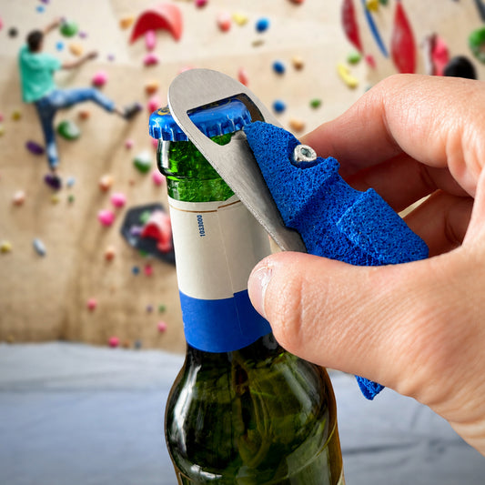 A close-up of a hand using a blue textured climbing hold bottle opener tool to remove a green bottle cap from a beer bottle, with a blurred climbing wall in the background.