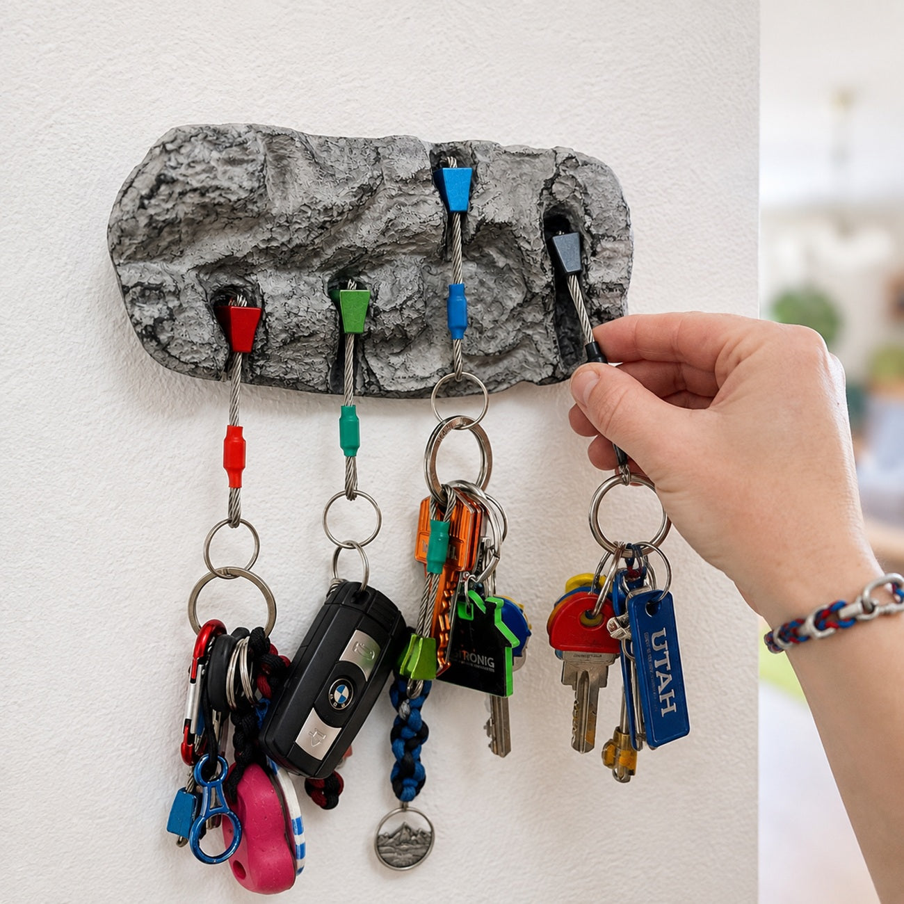 A textured gray rock-shaped key holder mounted on a white wall, holding several keys on metal rings. A person's hand is reaching in to adjust or hang a key.