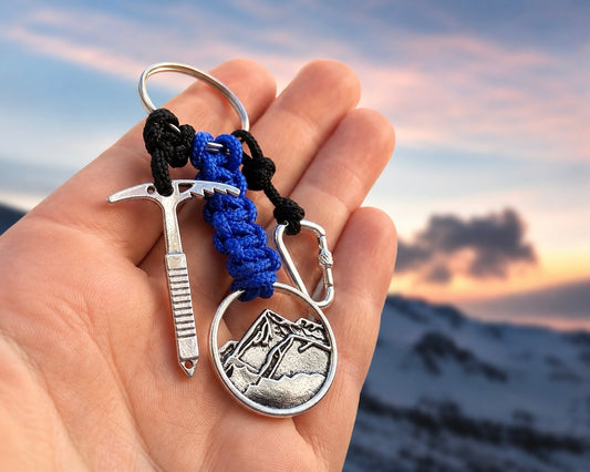 A hand holding a silver-toned mountain climbing keychain with a pickaxe, carabiner, and round medallion charm, attached to black and blue paracord, against a blurred mountain sunset background.