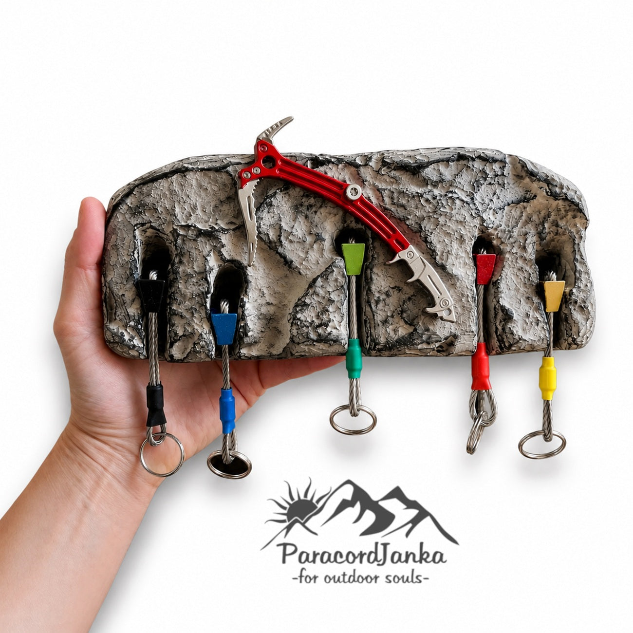 A hand holds a rock-climbing gear organizer shaped like a textured boulder, with five colored hooks holding climbing tools and keys. A red climbing axe is placed on top.
