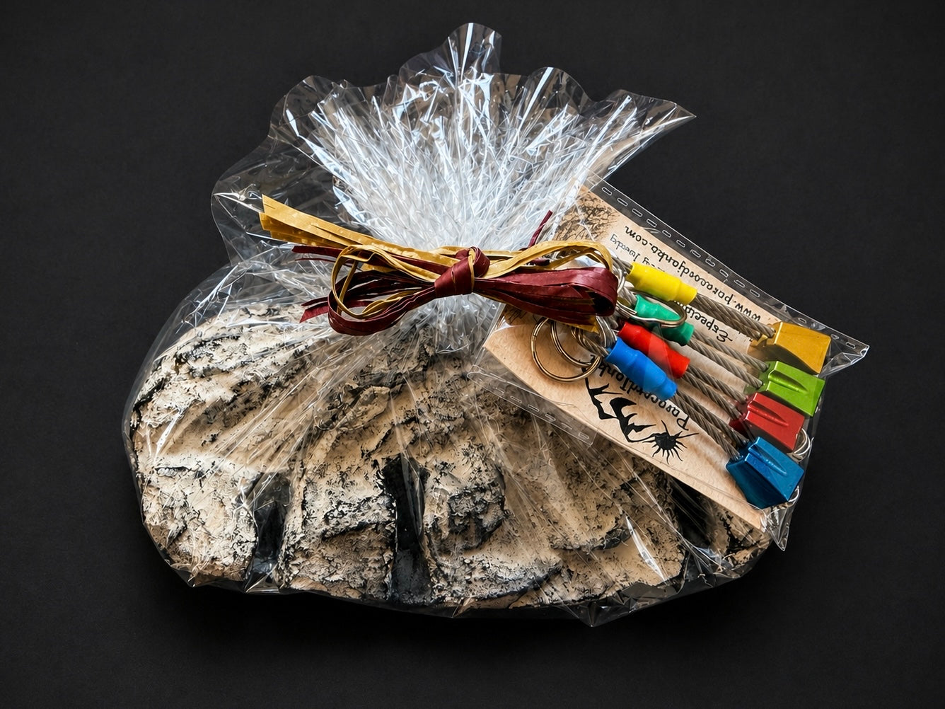 A gift set in clear cellophane wrapping featuring a large textured stone, a bundle of tan leather cords, and a small clear plastic package of colorful wooden keychains on a black background.
