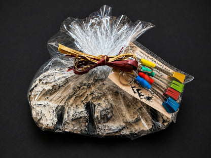 A gift set in clear cellophane wrapping featuring a large textured stone, a bundle of tan leather cords, and a small clear plastic package of colorful wooden keychains on a black background.
