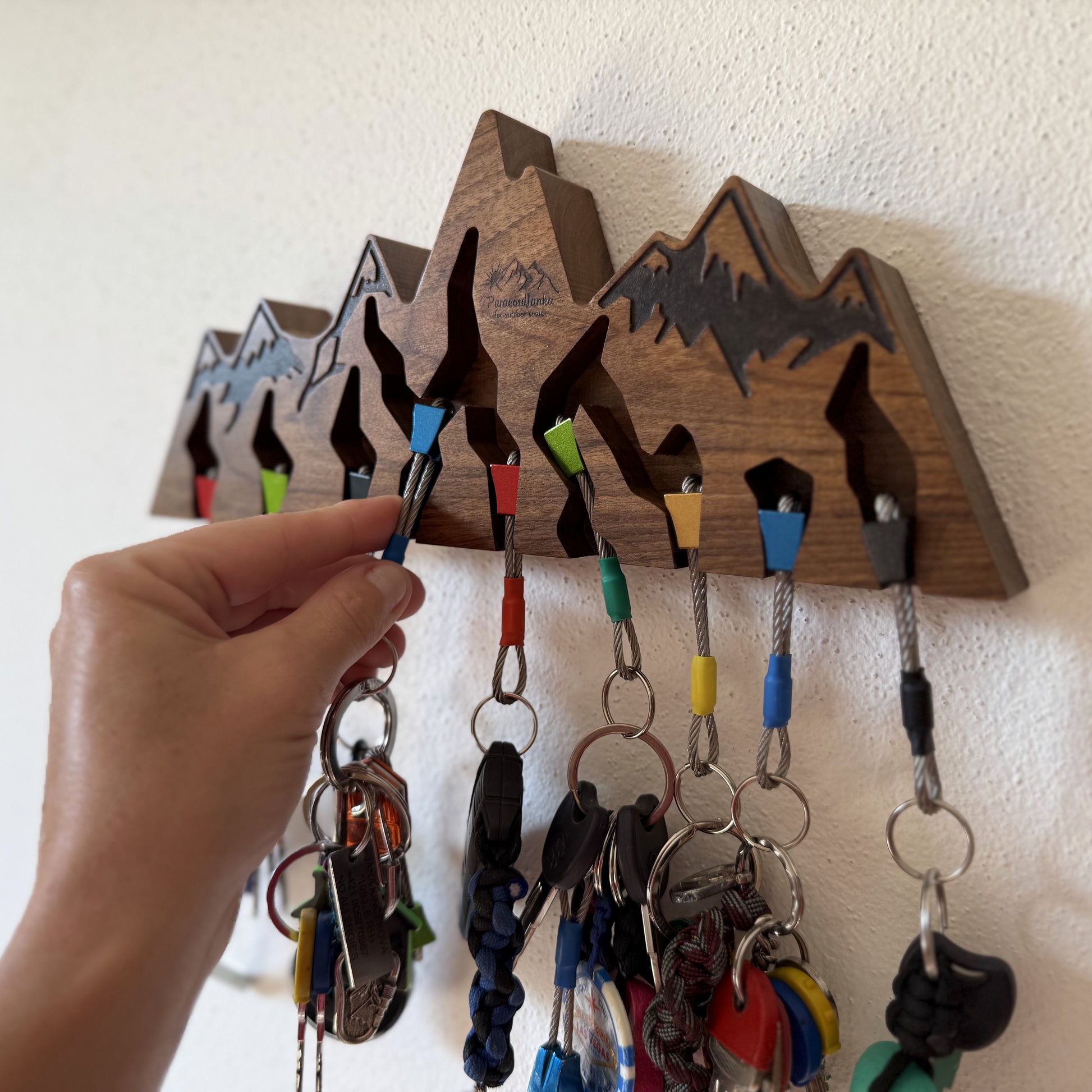 A wooden mountain-shaped key holder mounted on a white wall, with several colorful rope lanyards and keys hanging from its slots. A hand is reaching in to adjust or hang a key, showing use in a real interior setting.