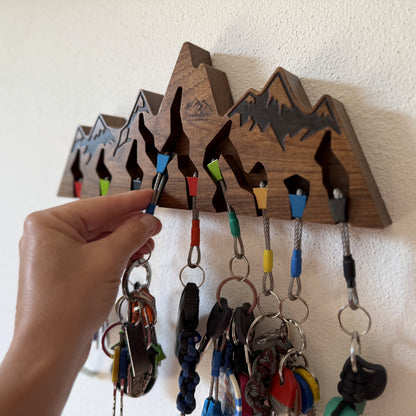 A wooden mountain-shaped key holder mounted on a white wall, with several colorful rope lanyards and keys hanging from its slots. A hand is reaching in to adjust or hang a key, showing use in a real interior setting.
