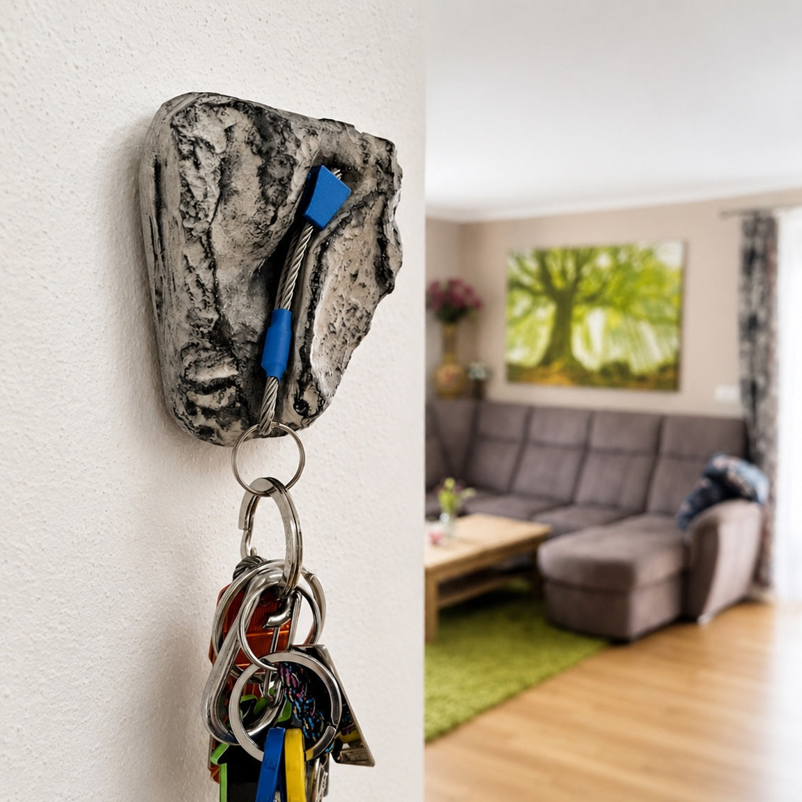 A rock-shaped key holder mounted on a white wall, holding a set of keys with blue and yellow keychains. The background shows a blurred living room with a gray sofa and wooden floor.