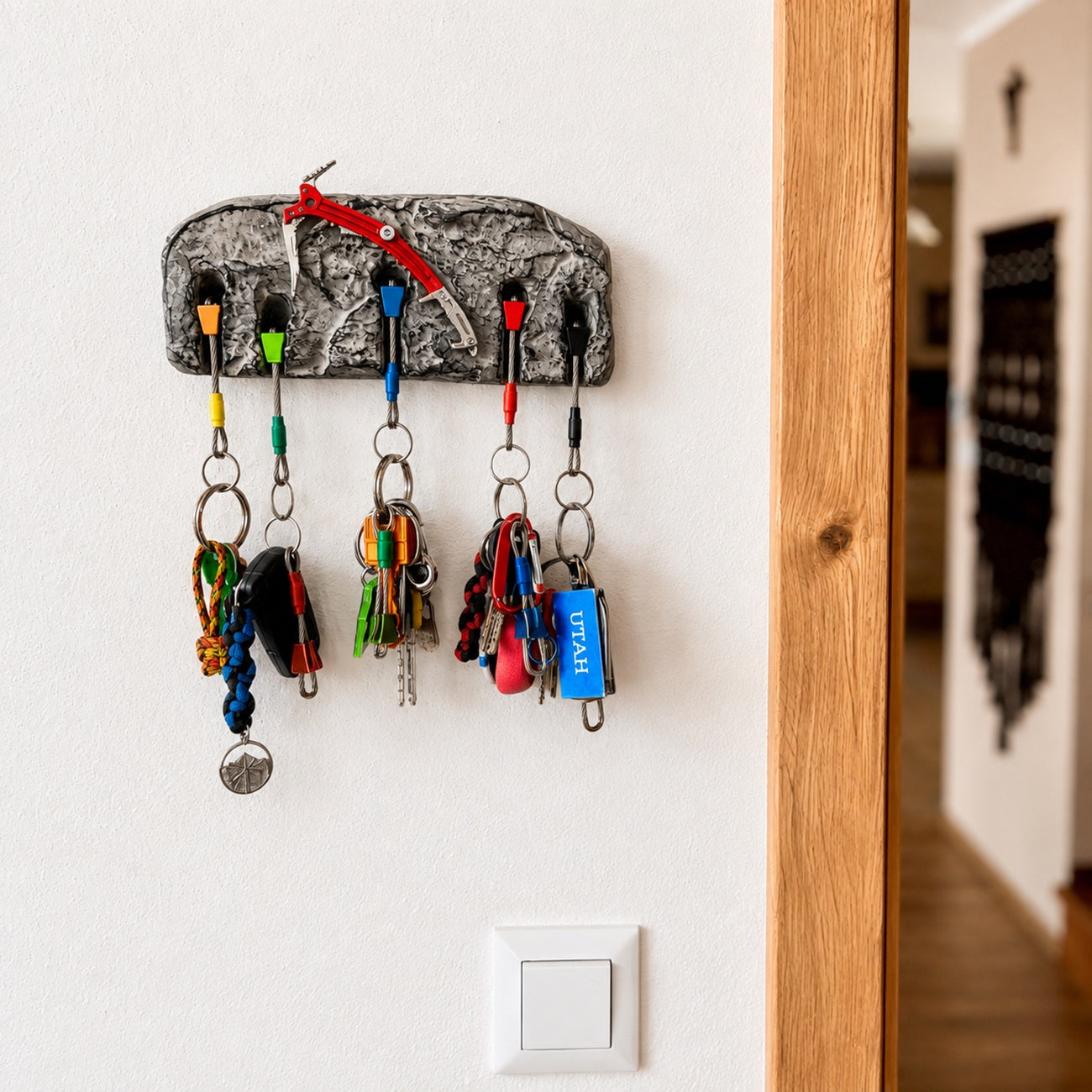A textured gray and black bark-style key holder mounted on a white wall, holding several colorful keys and keychains. A wooden door frame is visible on the right, and a light switch is below the holder.