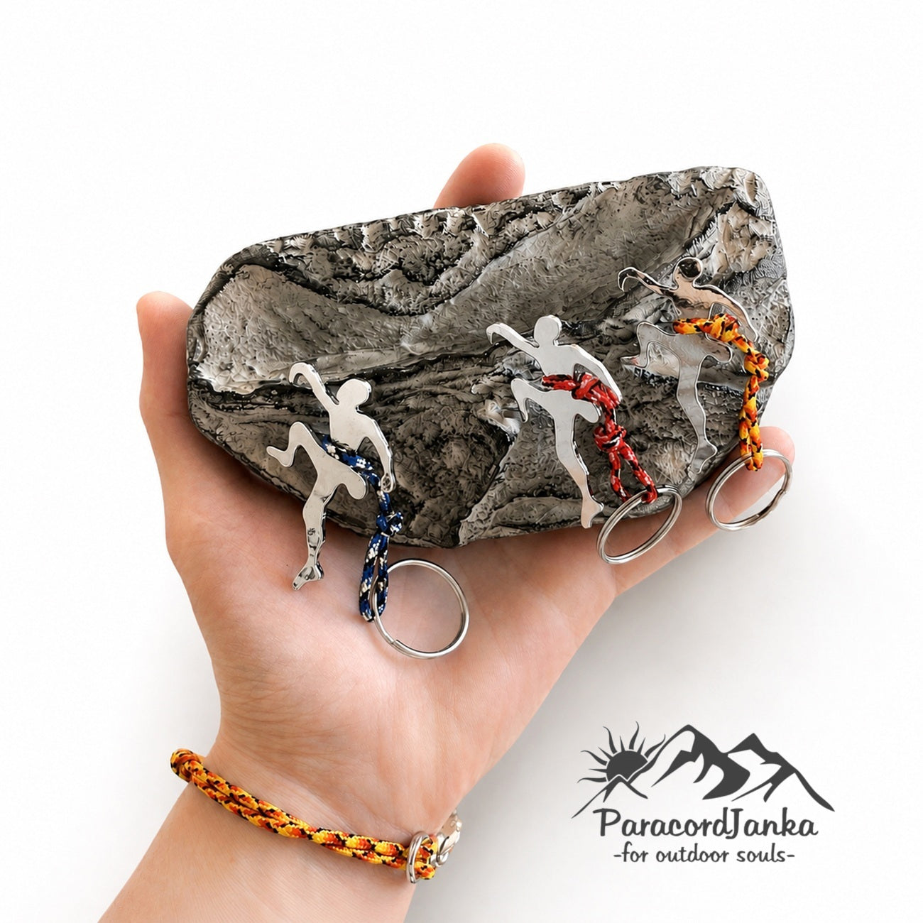 A hand holds a textured gray rock displaying three different colored outdoor-themed keychains. The keychains feature silver metal hikers and climbers with braided cords in blue, red, orange, and yellow.