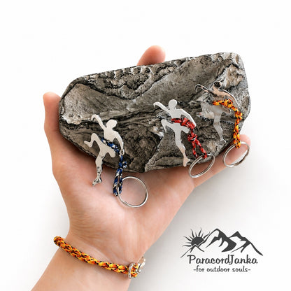 A hand holds a textured gray rock displaying three different colored outdoor-themed keychains. The keychains feature silver metal hikers and climbers with braided cords in blue, red, orange, and yellow.