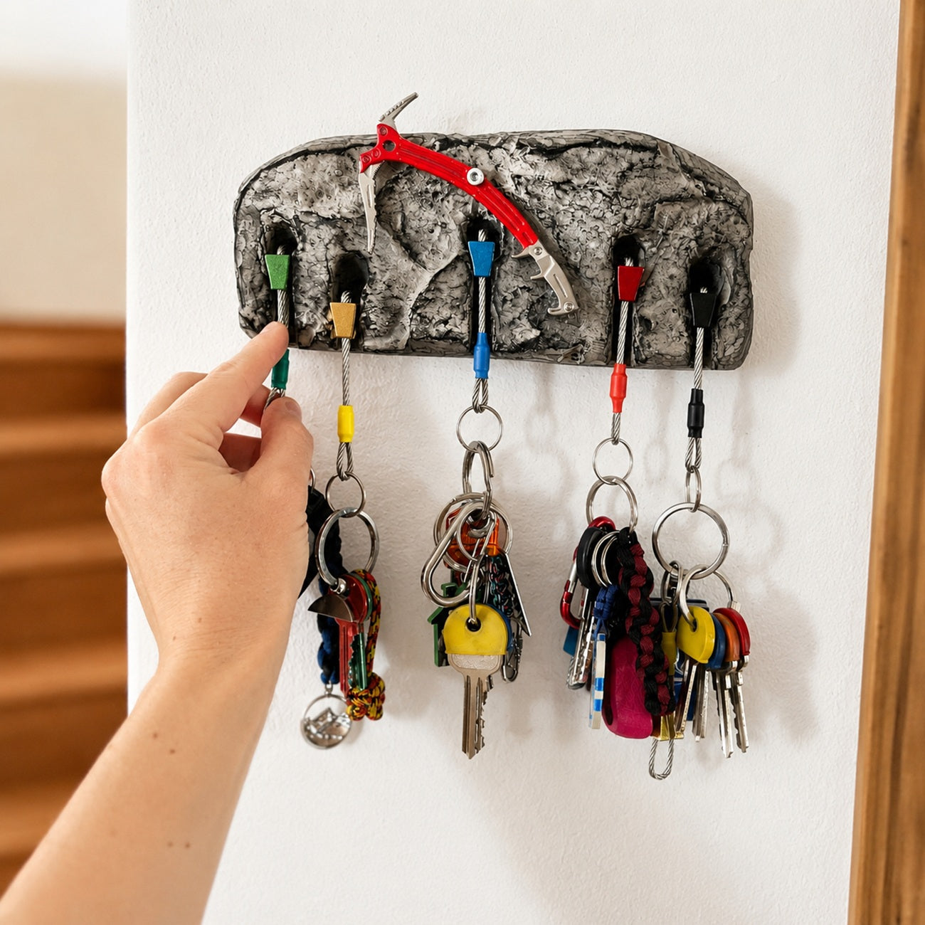 A hand holding a green key ring in front of a gray rock-themed key holder with climbing gear hooks and colorful keys hanging from it.
