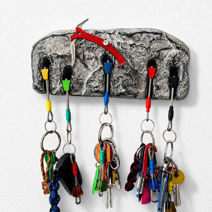 A novelty key holder shaped like a textured gray rock, featuring six colorful metal key hooks and several keys hanging from them, with a red climbing harness detail at the top, shown against a plain white wall.