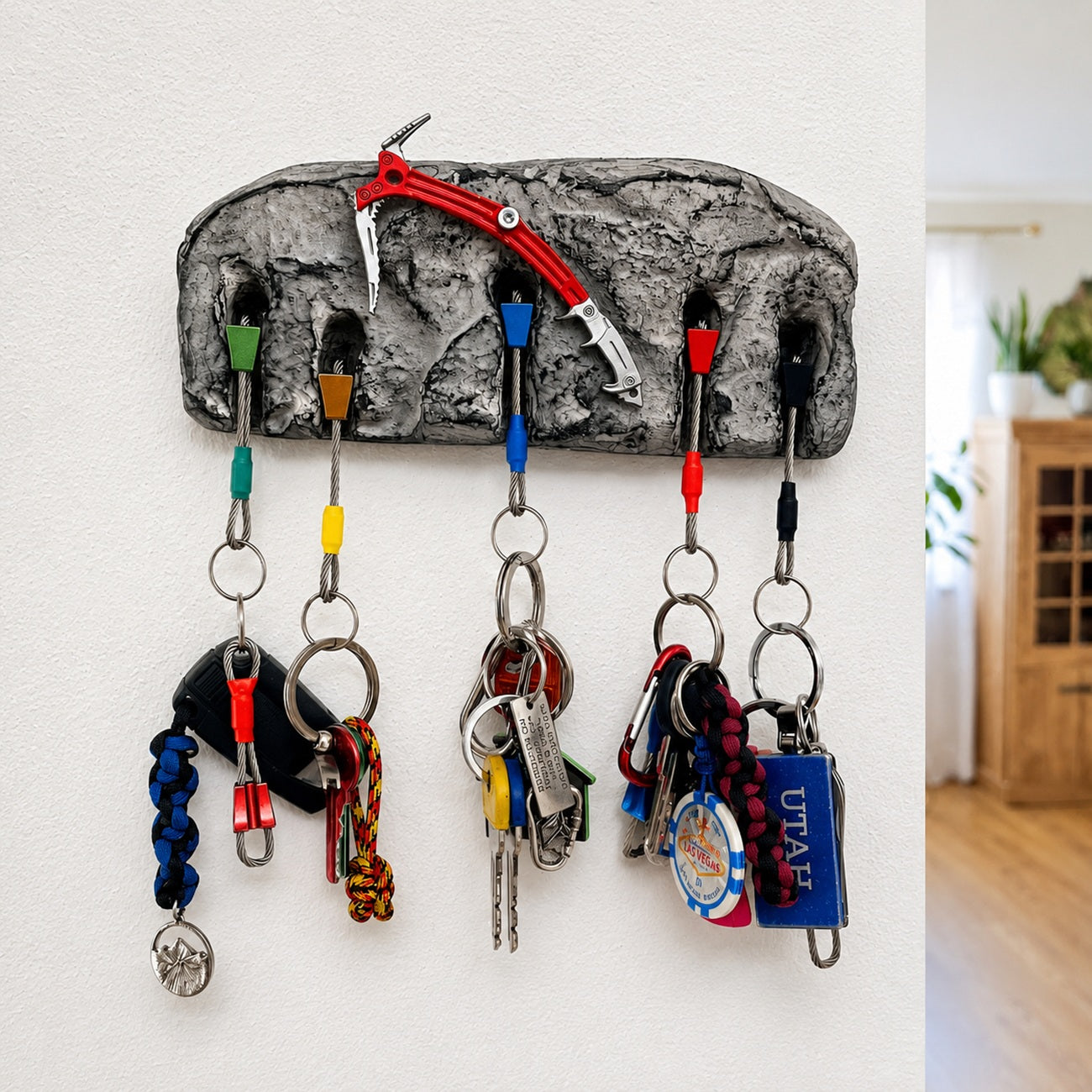 A gray rock-shaped wall key holder with five hooks holding various keys and keychains, shown mounted on a white wall with a glimpse of a home interior on the right.