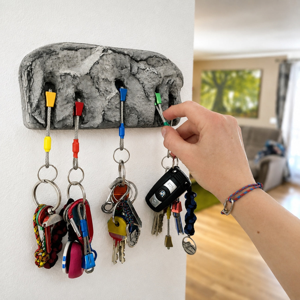A gray rock-textured wall-mounted key rack with five colorful hooks holding various keys and car keys, shown in a real indoor room with a hand reaching for one key.