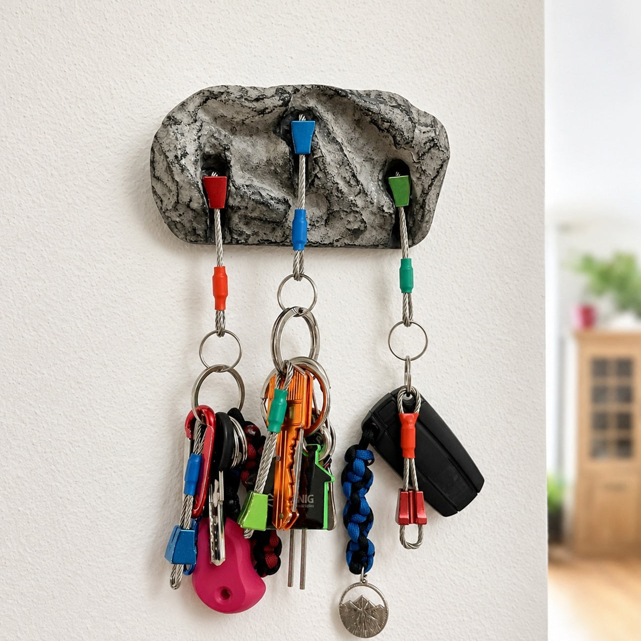 A rock-shaped wall key holder with three colorful hooks holding various keys and keychains, mounted on a white wall.