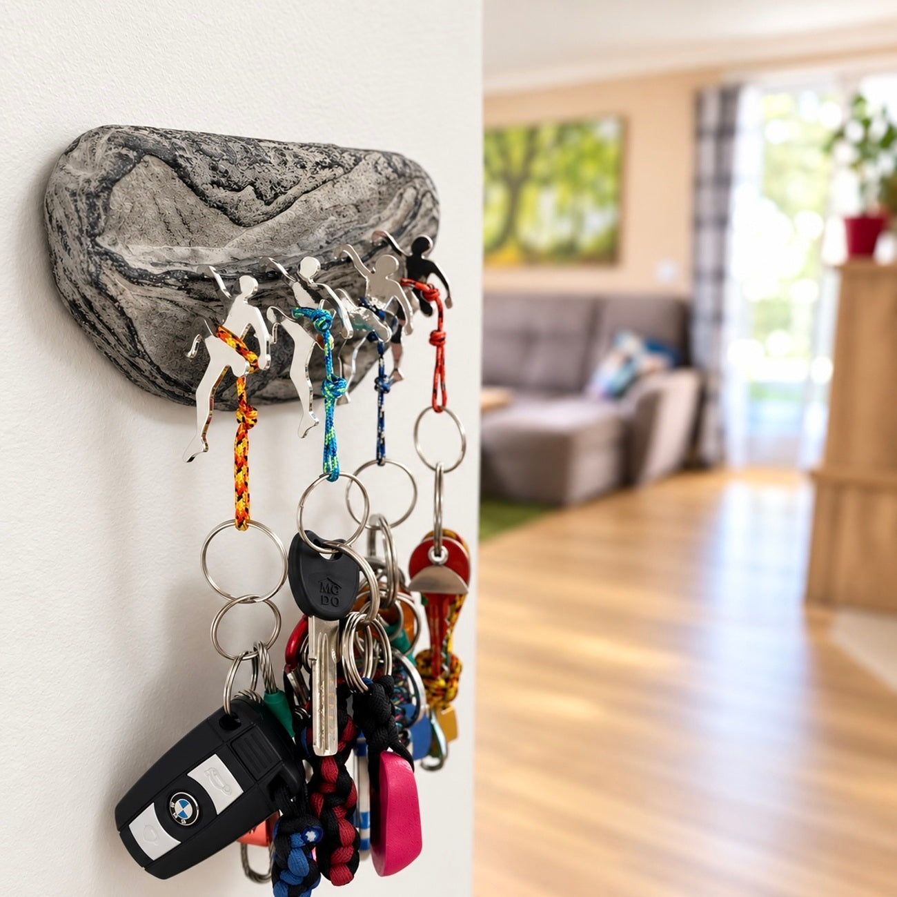 A rock-shaped wall hook holder mounted on a white wall, holding several colorful keychains and keys in a real indoor living room setting.