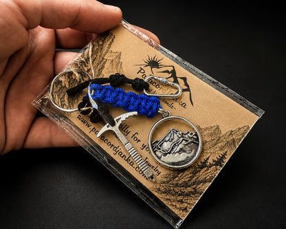 A hand holds a clear plastic package containing a small jewelry set with a mountain pendant, a silver-toned tool-shaped charm, and two macrame keychains in black and blue, all mounted on a kraft paper card with engraved mountain illustrations.