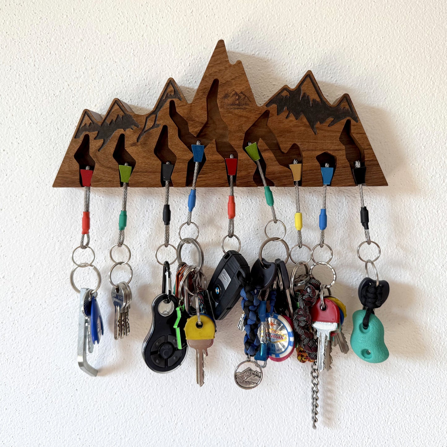 A wooden mountain-shaped key holder with multiple colorful hanging keys and keychains, mounted on a white textured wall.