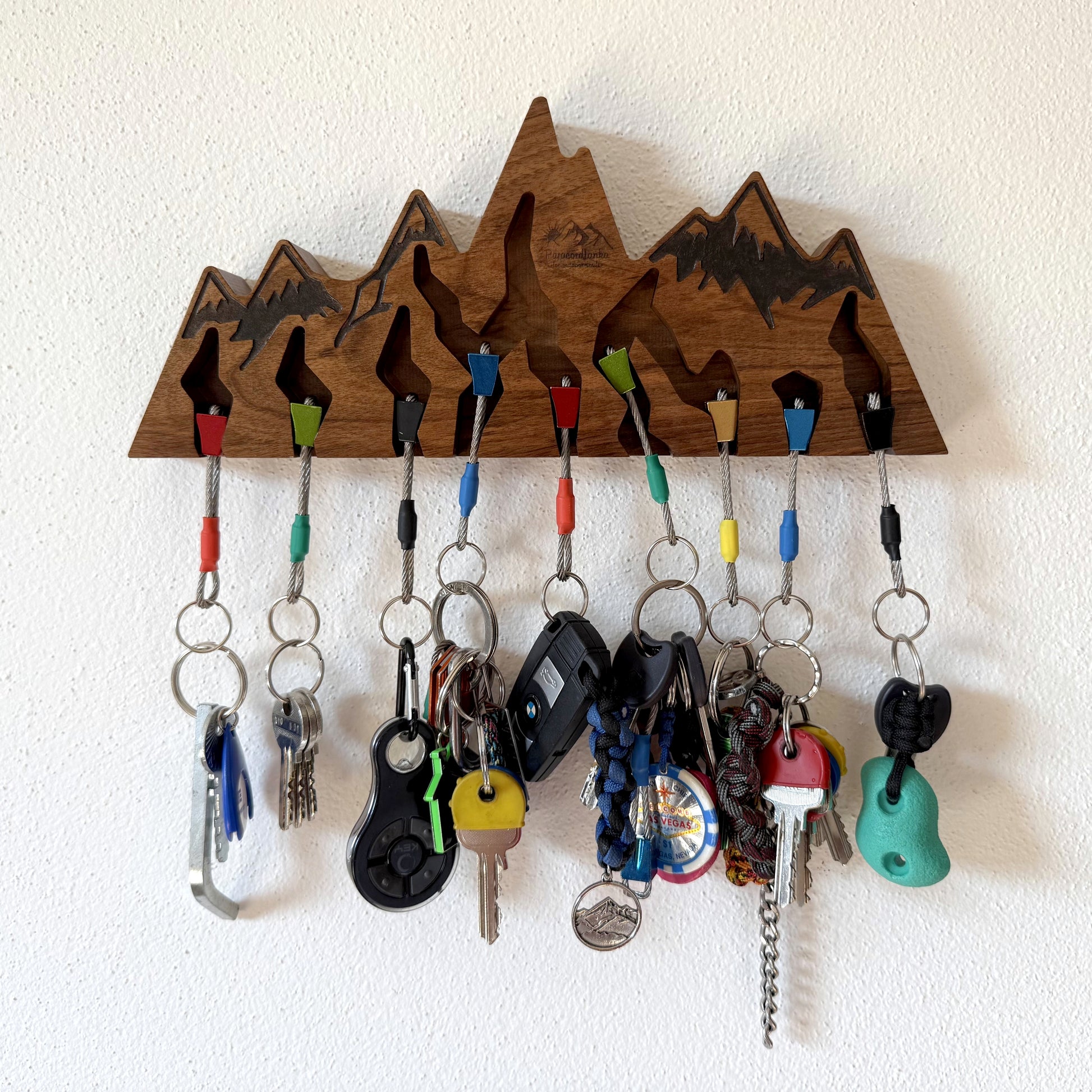 A wooden mountain-shaped key holder with multiple colorful hanging keys and keychains, mounted on a white textured wall.