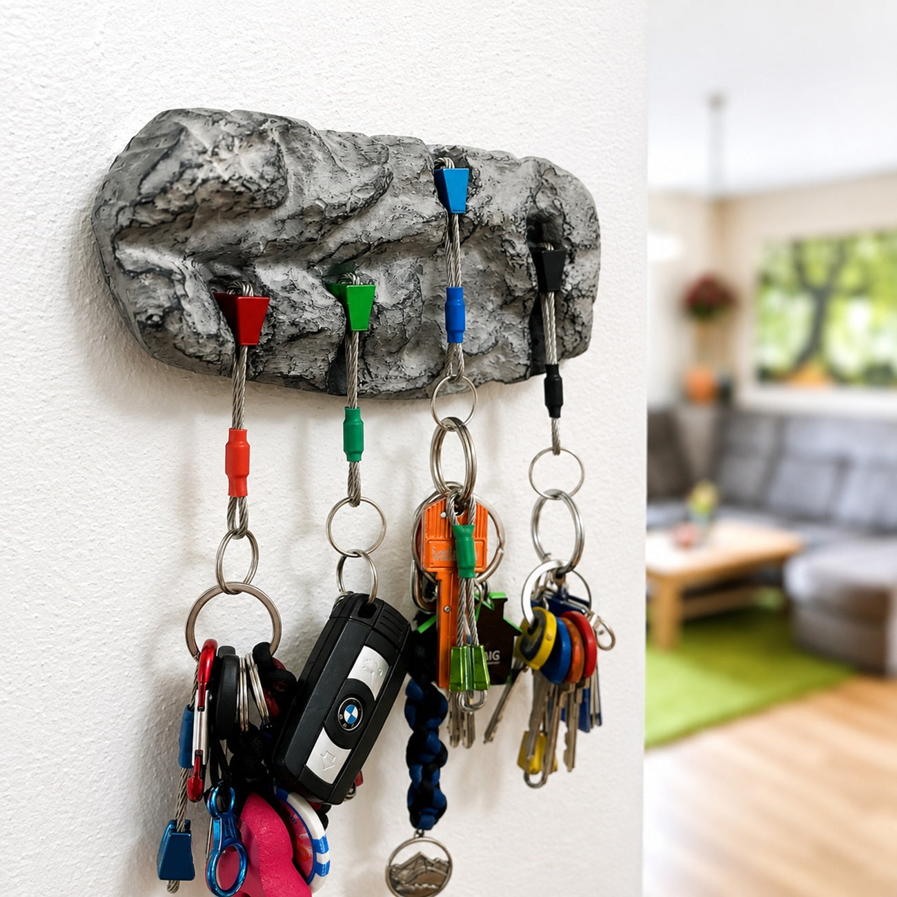 A rock-shaped wall key holder with five hooks, shown mounted on a white wall in a real living room. The holder has colorful keychains and keys hanging from it, including a BMW key fob.