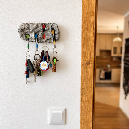A textured gray stone wall hook holder mounted on a white wall, holding several colorful keys and car keys, with a blurred kitchen visible in the background through a doorway.