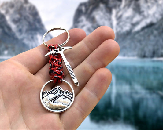 A silver keychain with a red and black braided cord. The keychain features a small ice axe charm and a round charm with a mountain silhouette.