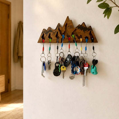 A wooden mountain-shaped key rack mounted on a white wall, holding multiple colorful keys and keychains in a home interior setting.