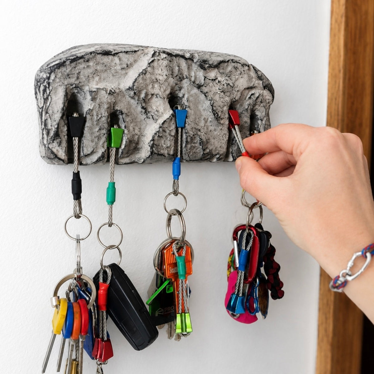 A textured gray key holder with five hooks mounted on a white wall, holding several colorful keychains and keys. A person's hand is shown reaching for one of the keys.