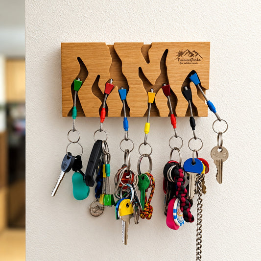 A wooden wall-mounted key rack with cutout shapes holding multiple colorful keychains and keys, mounted on a white wall. The wood has an engraved logo reading 'MountainLofts keyranging wall' in the top right.